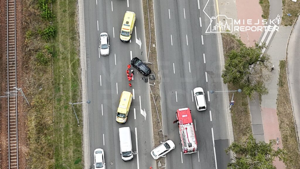 Na jezdni leży czarny samochód, a ratownicy udzielają pomocy poszkodowanemu; w tle widoczne inne pojazdy.
