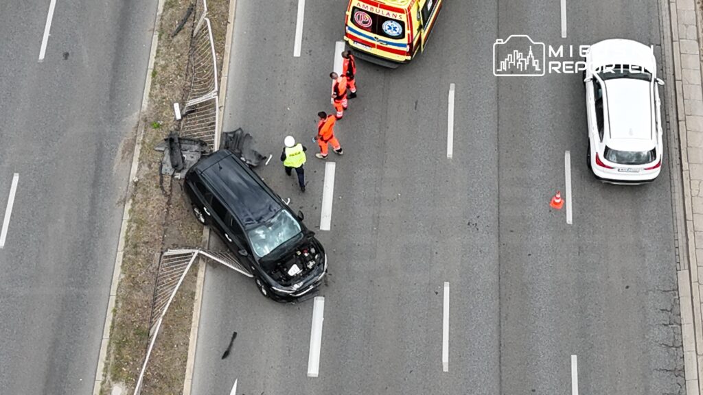 Czarny samochód osobowy stoi na poboczu drogi, obok grupy trzech ratowników w pomarańczowych uniformach i policjanta.