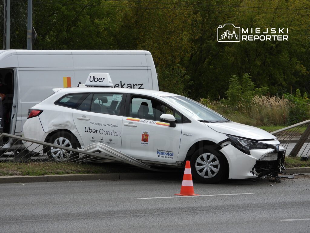 Biały samochód osobowy z logo Ubera wjeżdża w barierkę na poboczu drogi, obok stoi dostawczy pojazd.