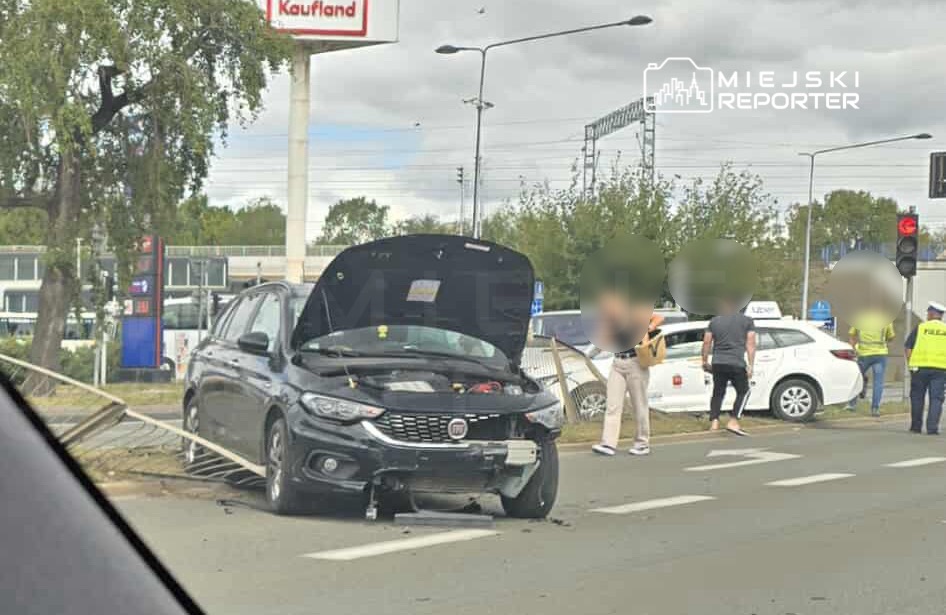 Czarny samochód osobowy z otwartą maską stoi na poboczu drogi, obok grupy osób rozmawiających z policjantem.