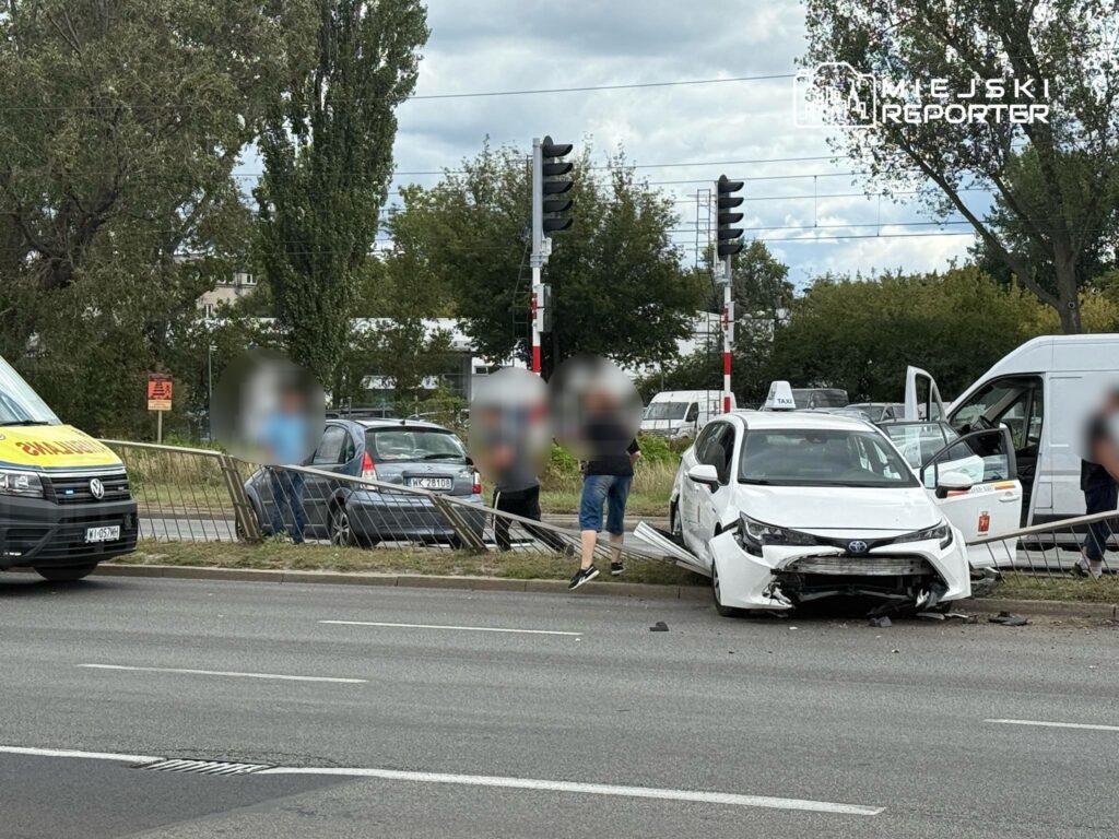 W wyniku kolizji samochodów, biała taksówka z uszkodzoną przednią częścią stoi przy ogrodzeniu, obok niej grupa osób.