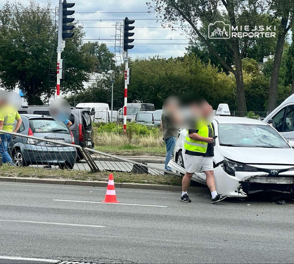 Na skrzyżowaniu z sygnalizacją świetlną, mężczyzna w kamizelce odblaskowej ocenia uszkodzenia białego samochodu po kolizji.