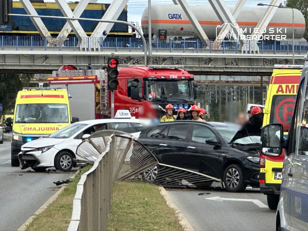 W miejscu wypadku drogowego kilka służb ratunkowych udziela pomocy poszkodowanym, a na jezdni leżą uszkodzone pojazdy.