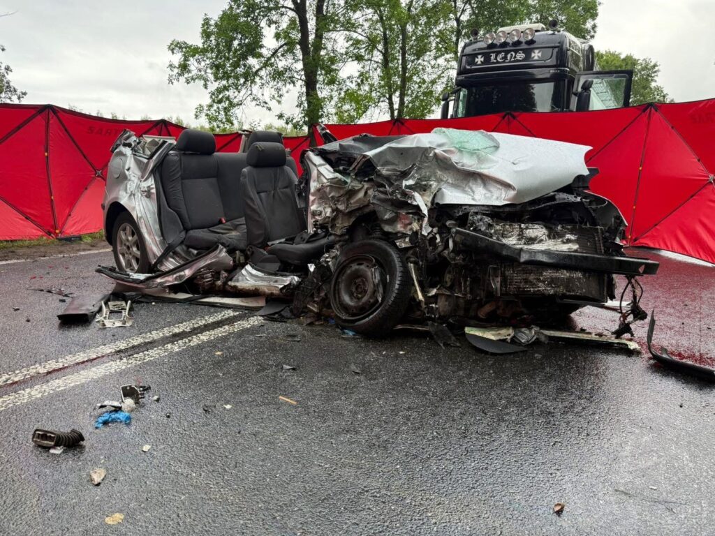 Zniszczony samochód osobowy leży na drodze, otoczony przez czerwone namioty, obok stoi ciężarówka.
