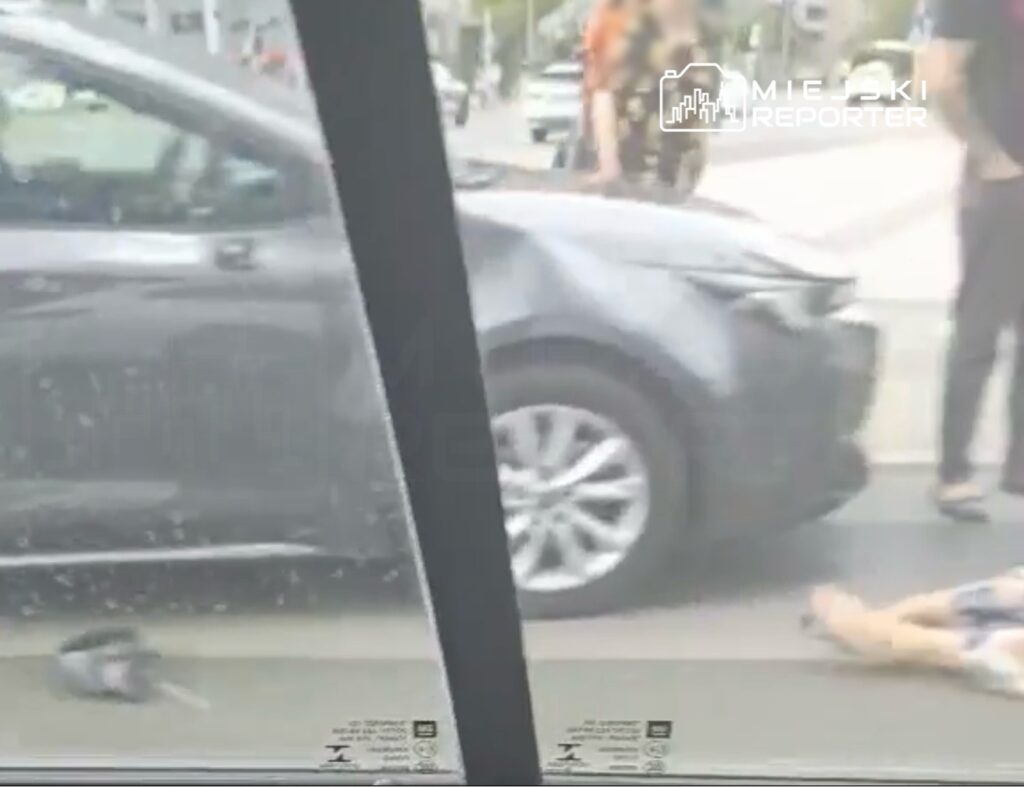 Wypadek drogowy z osobą leżącą na jezdni obok uszkodzonego samochodu, w otoczeniu przechodniów.