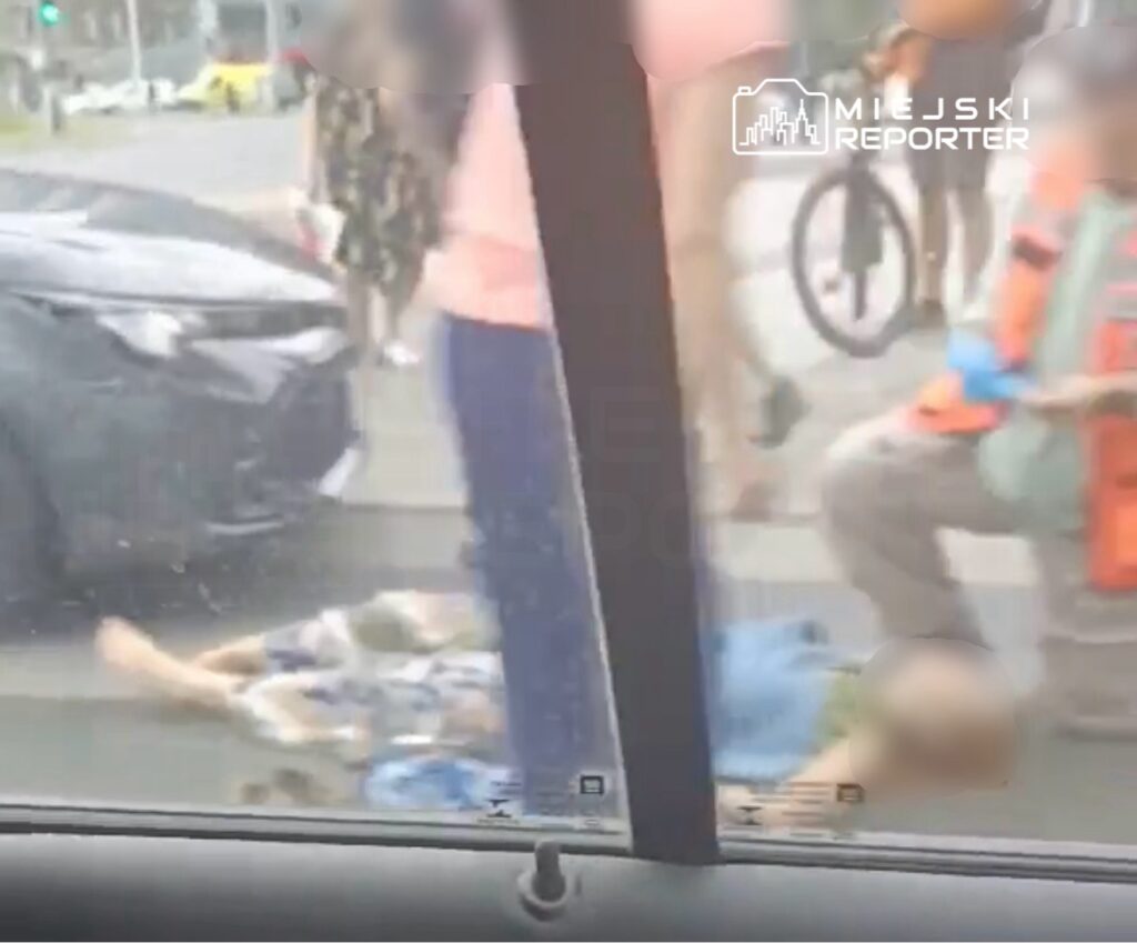 Na jezdni leży osoba, obok niej stoją przechodnie oraz pracownik w pomarańczowej kamizelce.
