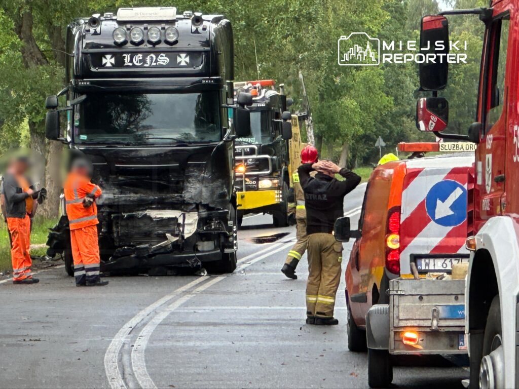 Czarny ciężarówka z uszkodzoną przednią częścią stoi na drodze, obok pracownicy w pomarańczowych ubraniach i strażak w hełmie.