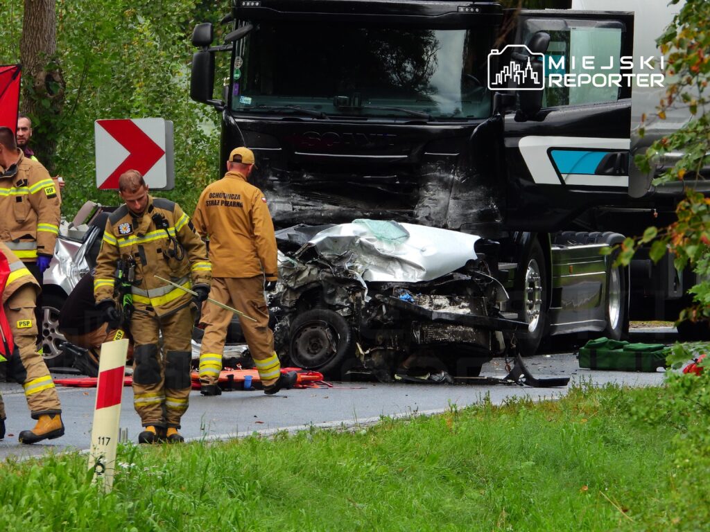 Grupa strażaków pracuje nad usunięciem wraku rozbitego samochodu osobowego po zderzeniu z ciężarówką na drodze.