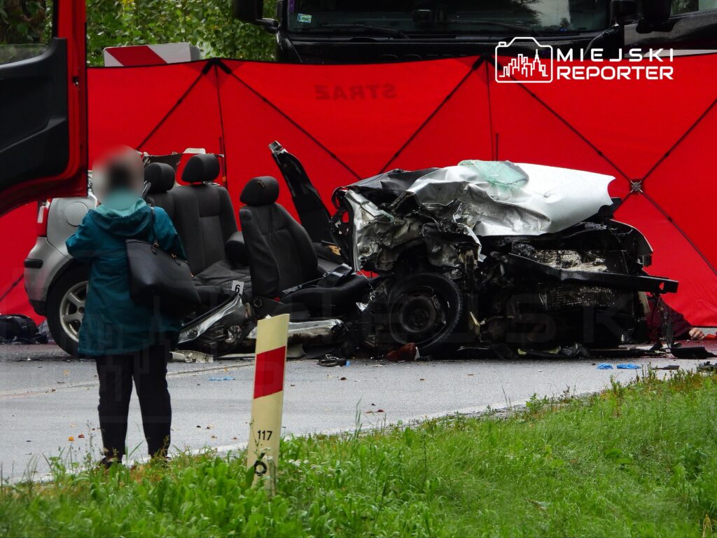 Kobieta w niebieskiej kurtce stoi obok zniszczonego samochodu osobowego, otoczonego czerwonymi ekranami ochronnymi.
