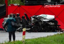 Kobieta w niebieskiej kurtce stoi obok zniszczonego samochodu osobowego, otoczonego czerwonymi ekranami ochronnymi.