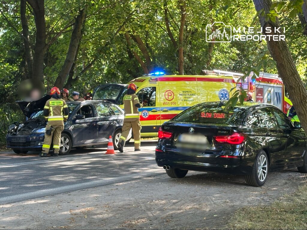 Grupa strażaków w mundurach pomaga przy wypadku drogowym, obok stoi ambulans i czarny samochód osobowy.