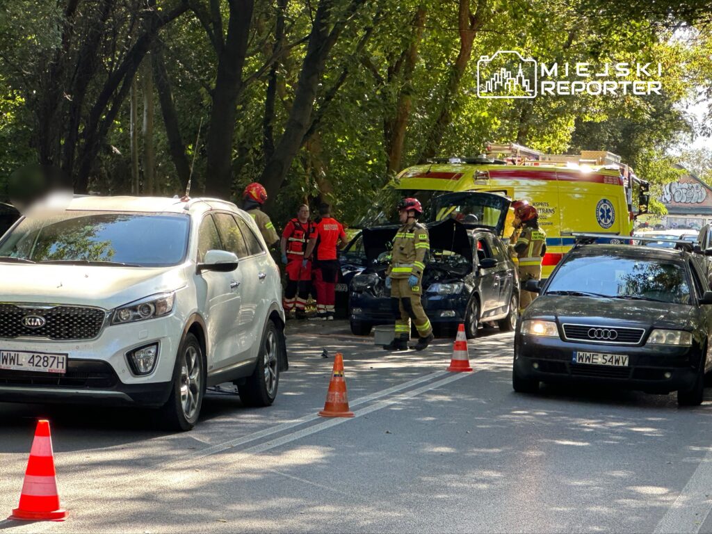Na drodze stoją dwa samochody, w tym biały SUV, obok których pracują strażacy w hełmach. W tle widoczna karetka pogotowia.
