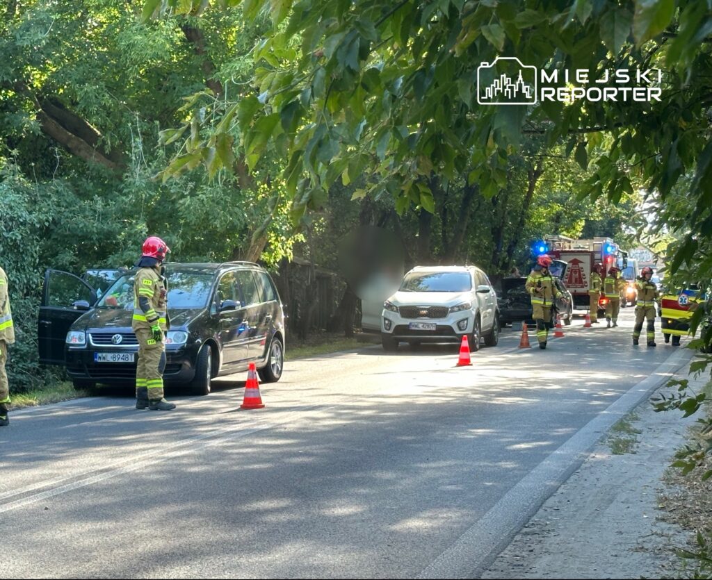 Strażacy w kaskach i mundurach pracują przy czarnym samochodzie zaparkowanym na poboczu drogi, obok wozu strażackiego.