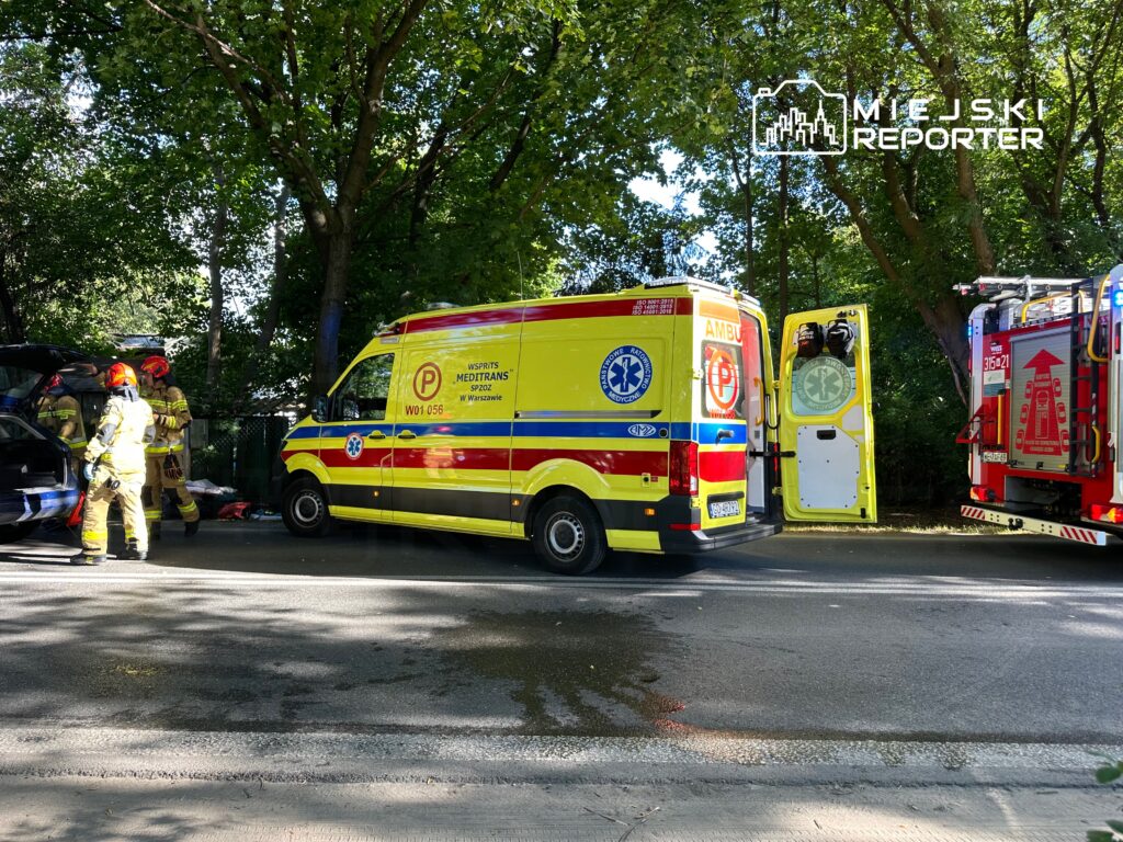 Ratownicy medyczni i strażacy pracują przy wypadku drogowym, obok dwóch pojazdów ratunkowych na zadrzewionej ulicy.