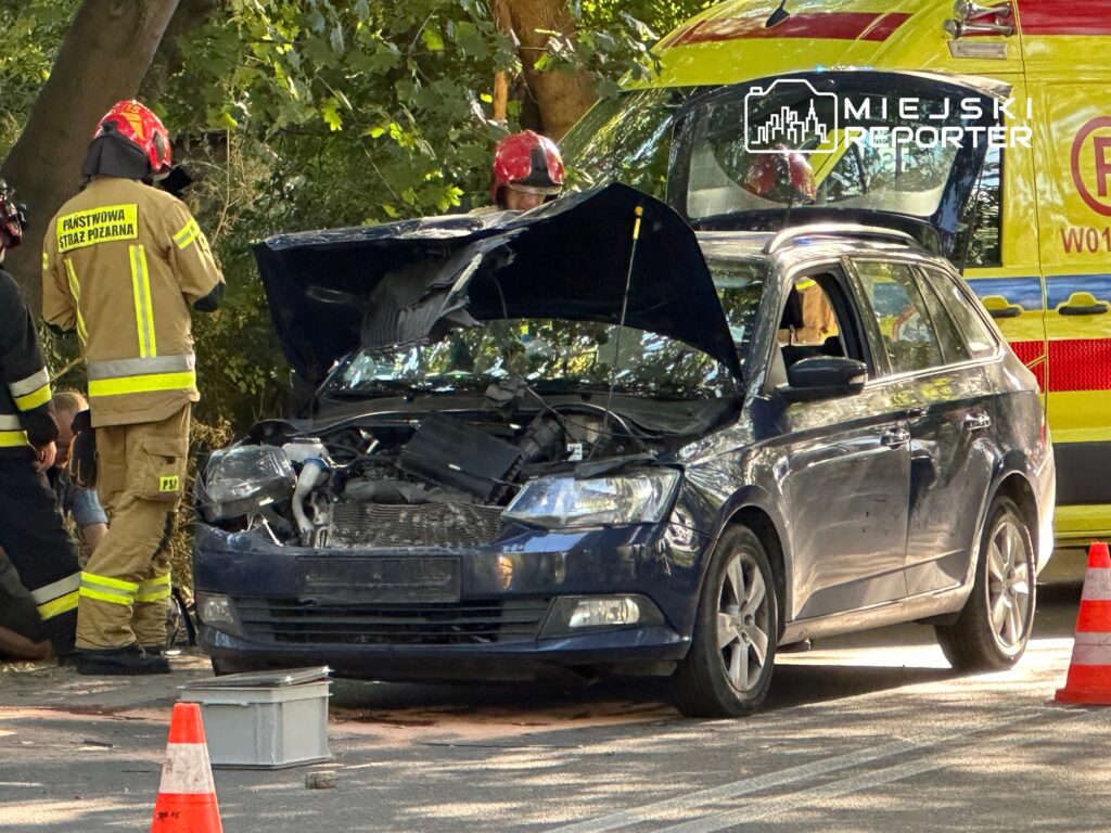 Strażacy w mundurach pracują przy uszkodzonym niebieskim samochodzie osobowym z otwartą maską, obok stoi ambulans.