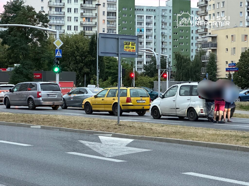 Grupa osób gromadzi się wokół białego samochodu na ulicy, w tle widoczne ruchliwe skrzyżowanie i budynki mieszkalne.