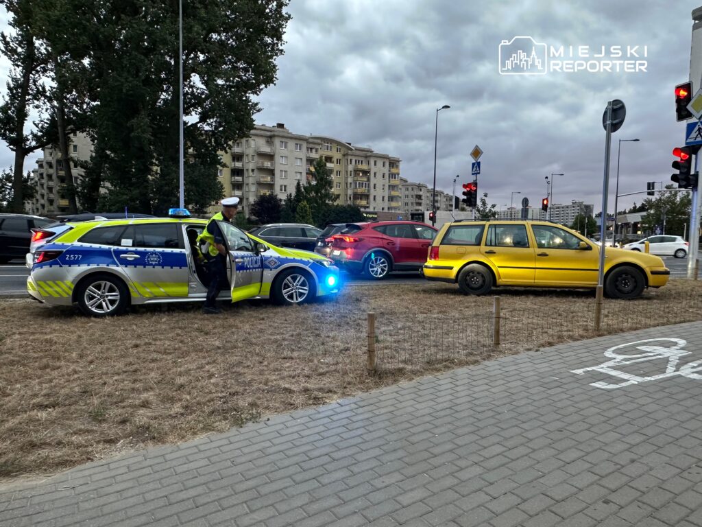 Policjant przy radiowozie z włączonymi sygnałami świetlnymi kontroluje sytuację na skrzyżowaniu, obok stoi żółty samochód.