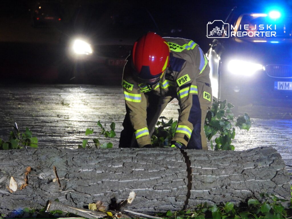 Strażak w hełmie i mundurze z odblaskami usuwa powalony pień drzewa z drogi w nocy, przy włączonych sygnalizacjach świetlnych.