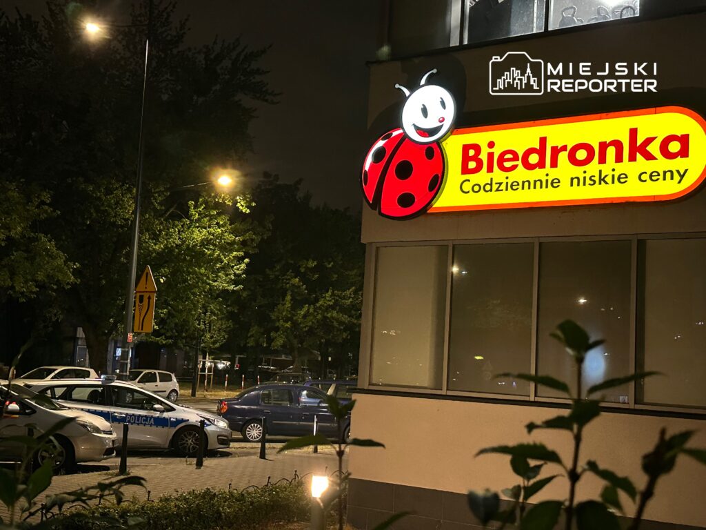 Oświetlona reklama sklepu Biedronka z hasłem "Codziennie niskie ceny" w otoczeniu zaparkowanych samochodów.