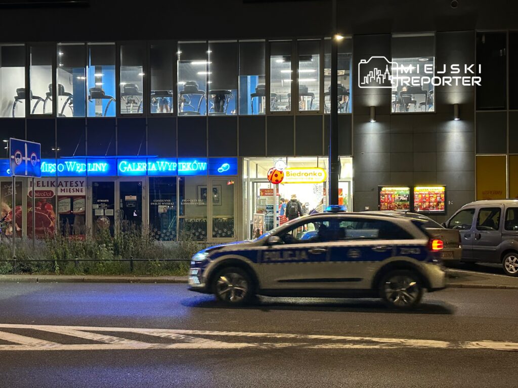 Policyjny radiowóz przejeżdża obok sklepu spożywczego Biedronka i lokalu gastronomicznego Galeria Wypieków.