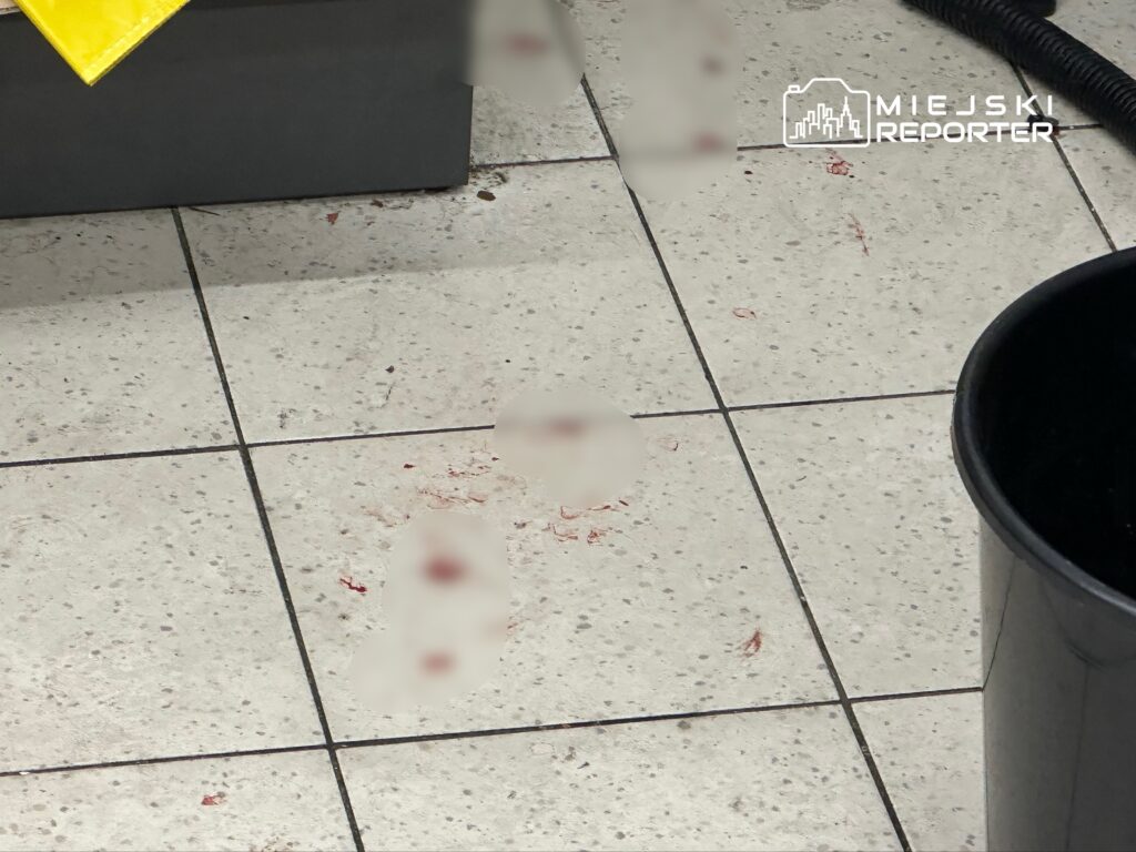 Plama krwi na kafelkowej podłodze w pomieszczeniu, obok czarny kosz na śmieci.