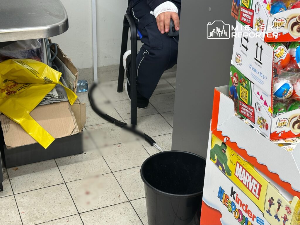 Mężczyzna w ciemnych spodniach siedzi na krześle obok stołu w sklepie, obok niego widoczna torba z gazetami i zabawkami.