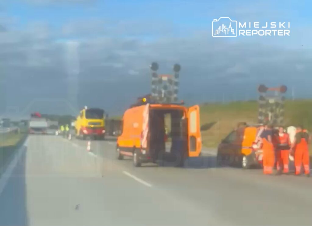 Ekipa ratunkowa w pomarańczowych strojach pracuje przy drodze, obok oznakowanych pojazdów i sygnalizacji świetlnej.