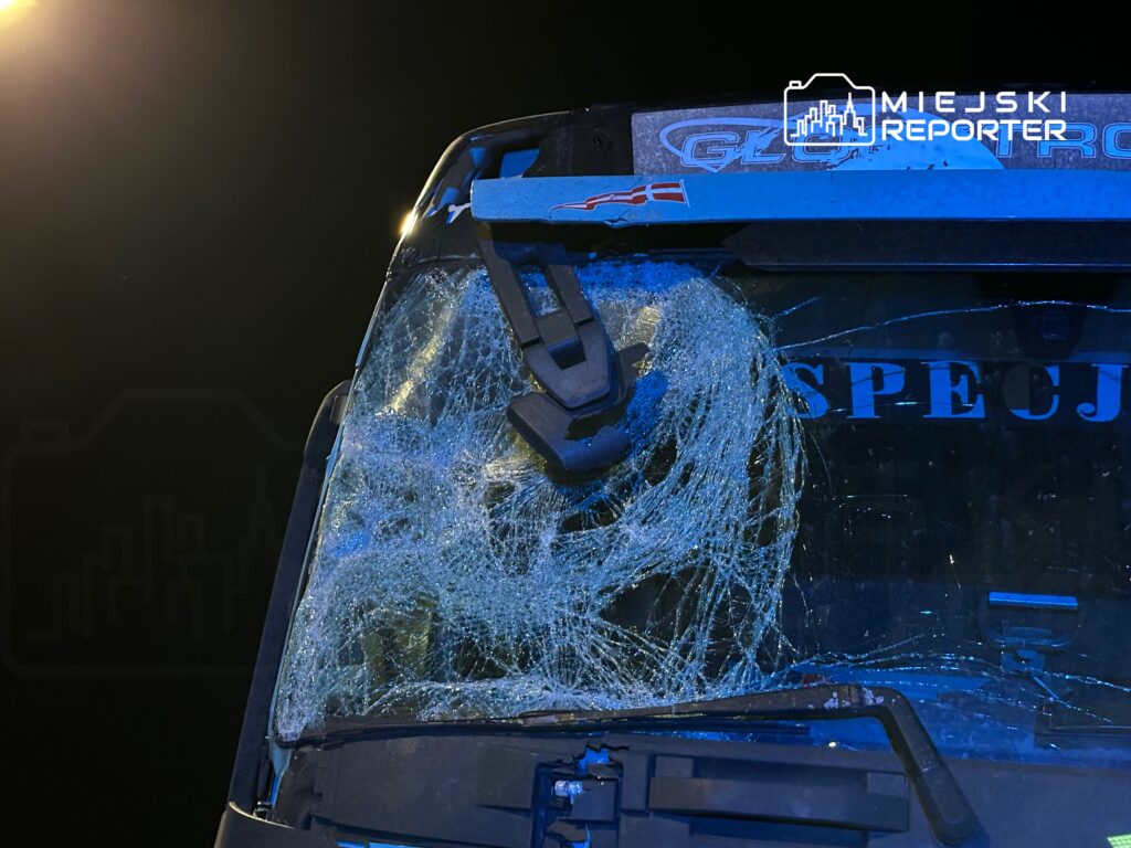 Zniszczona szyba czołowa pojazdu z pęknięciami, oświetlona przez lampy uliczne w ciemnej scenerii.
