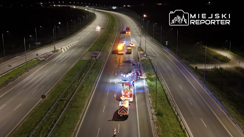 Na drodze nocą trwają działania służb ratunkowych, w tym samochody strażackie z włączonymi sygnałami świetlnymi.