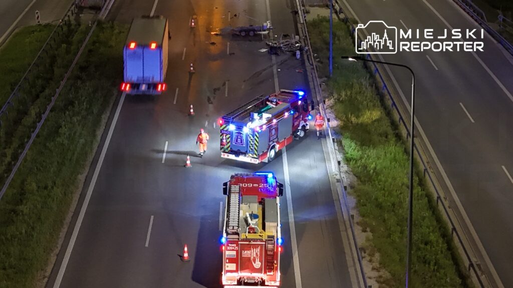 Na drodze stoją dwa wozy strażackie z włączonymi sygnałami świetlnymi, a obok pracują ratownicy.