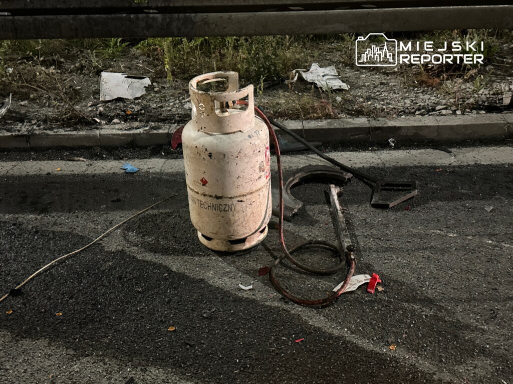 Butla gazowa stoi na asfalcie obok uszkodzonej metalowej rury i kawałków odpadków.