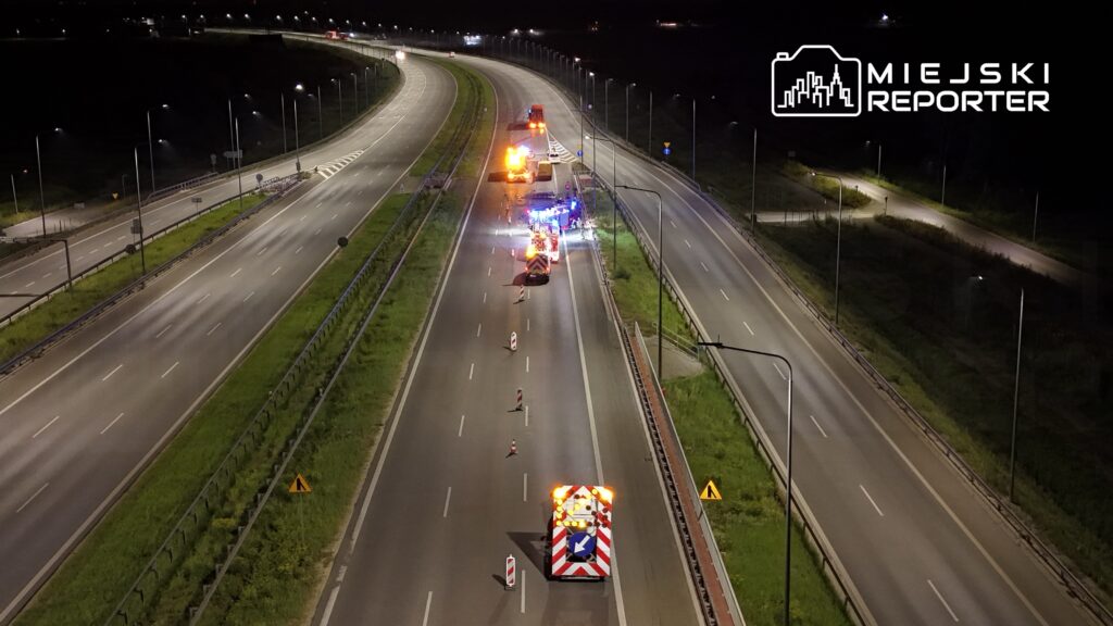 Na drodze nocą stoją pojazdy służb ratunkowych, a w tle widać oświetloną trasę z wyraźnymi znakami drogowymi.