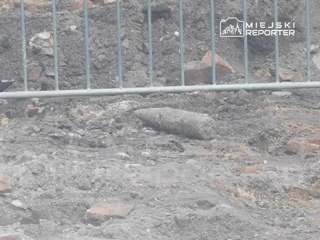 Na placu budowy widoczny jest pocisk leżący na ziemi, otoczony przez gruz i piasek, za ogrodzeniem z metalowych prętów.