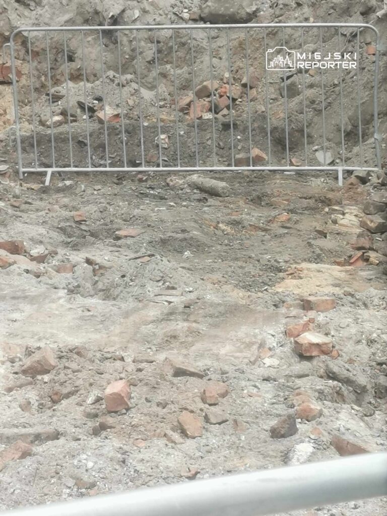 Plac budowy z ogrodzeniem, pokryty ziemią i fragmentami cegieł, z widocznymi śladami wykopów.