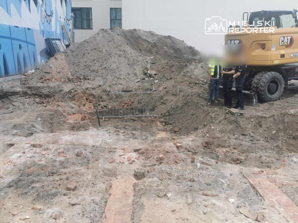Grupa osób w odzieży roboczej rozmawia obok wykopu, w tle widoczny pojazd budowlany oraz sterta ziemi i gruzu.