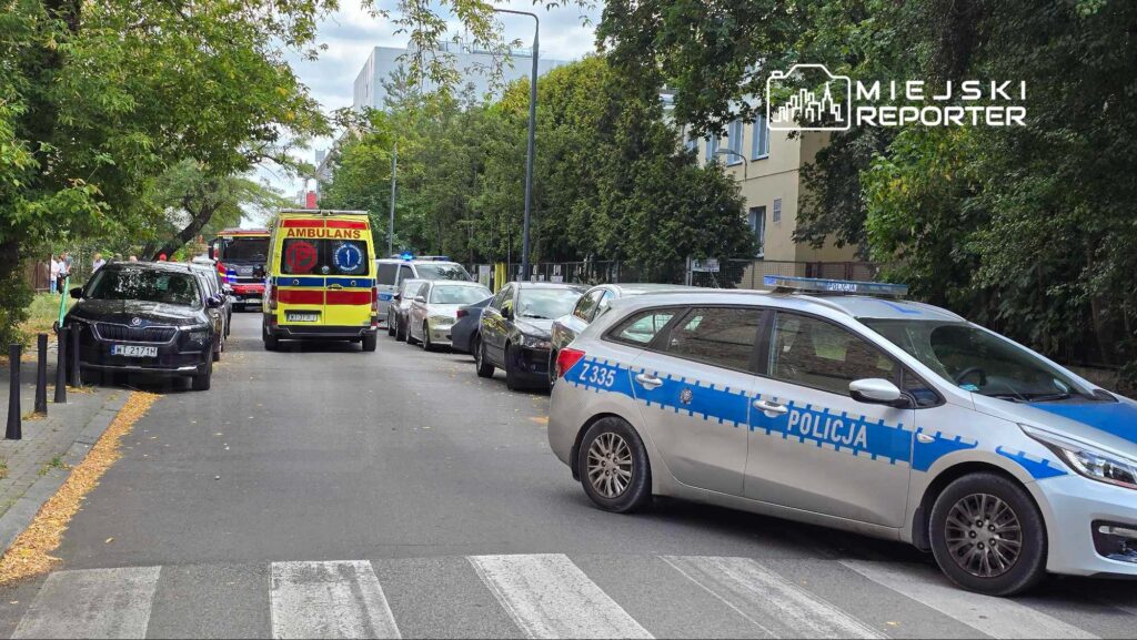 Karetka pogotowia oraz radiowóz policji zaparkowane na ulicy, w otoczeniu zaparkowanych samochodów i zieleni.