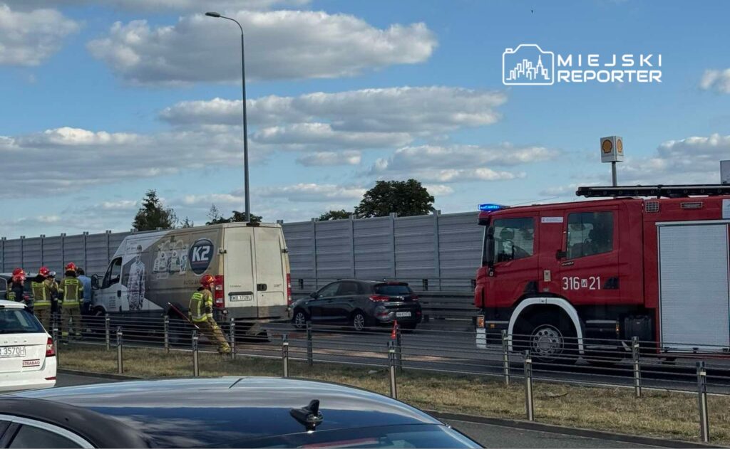 Grupa strażaków pracuje przy uszkodzonym pojeździe na pasie drogowym, obok stoi wóz strażacki.