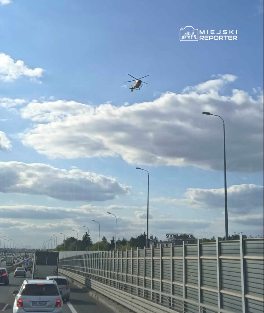 Helikopter unoszący się nad zatorowanym ruchem drogowym na autostradzie, w tle widoczne chmury.