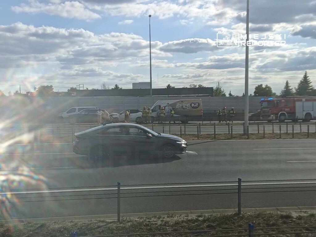 Na drodze autostradowej grupa osób w kamizelkach odblaskowych pracuje przy uszkodzonym pojeździe, obok stoi wóz strażacki.