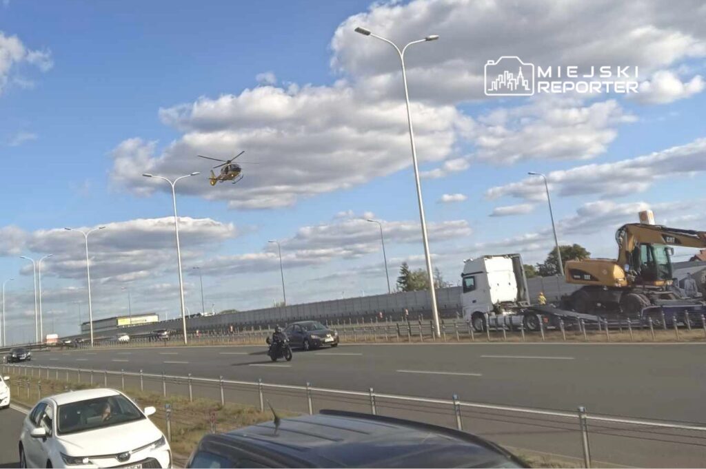 Helikopter unoszący się nad ruchem drogowym, w tle widoczne pojazdy i chmury na niebie.