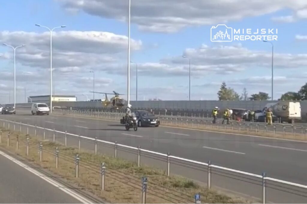 Na drodze widoczny jest policyjny motocykl oraz helikopter, w tle zespół ratunkowy pracujący przy wypadku.