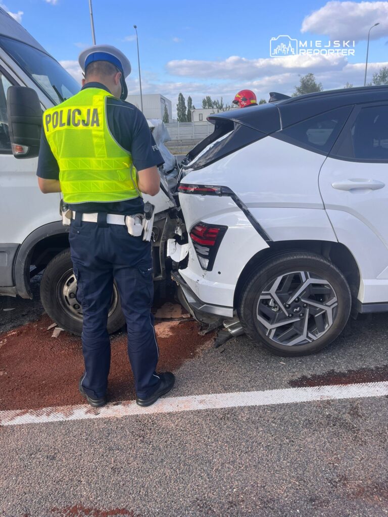 Policjant w kamizelce odblaskowej bada wypadek drogowy z udziałem białego SUV-a i furgonetki.