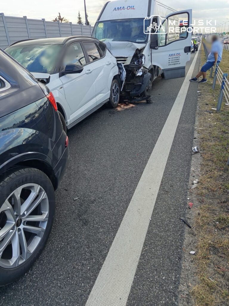 Zderzenie dwóch samochodów osobowych na drodze, jeden z pojazdów uszkodzony, w tle widoczny dostawczy samochód.