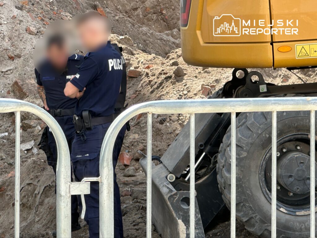Dwóch policjantów rozmawia obok żółtej koparki na placu budowy, otoczeni przez ziemię i gruz.