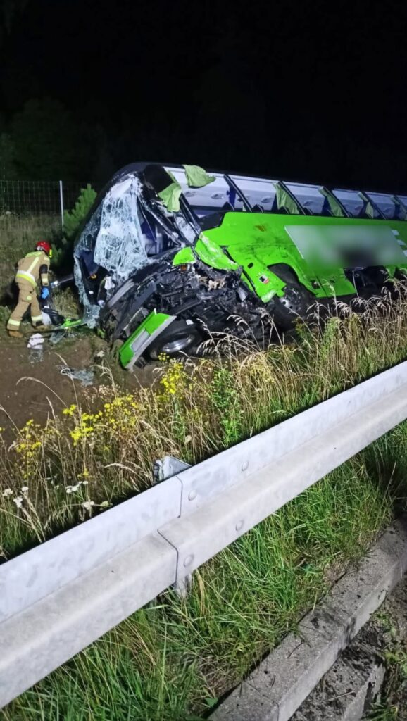 Zielony autobus przewrócony na poboczu drogi, obok strażak w mundurze przeszukuje wrak w nocy.