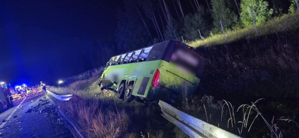Zielony autobus leży na poboczu drogi, obok grupy strażaków i pojazdów ratunkowych, w nocy, w lesistym otoczeniu.