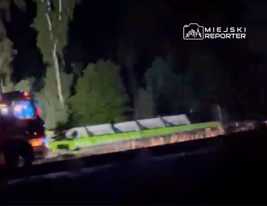 Czerwony wóz strażacki z włączonymi sygnałami świetlnymi stoi obok torów kolejowych w ciemnym otoczeniu.