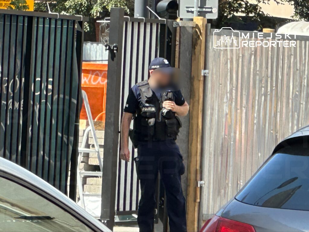 Policjant w mundurze stoi przy otwartych bramach, trzymając w ręku radiotelefon, w tle widoczne graffiti.