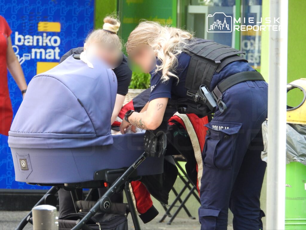Dwie policjantki sprawdzają zawartość wózka dziecięcego na ulicy, obok bankomatu.