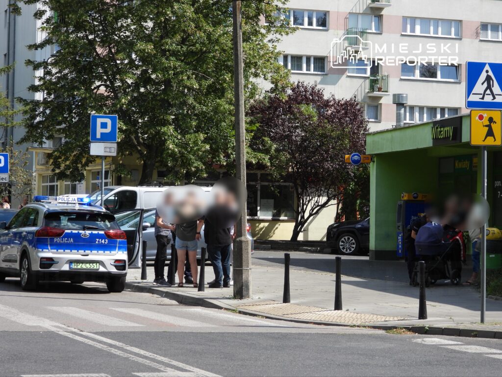 Grupa osób rozmawia z policjantami obok radiowozu na ulicy, w tle widoczny sklep spożywczy.